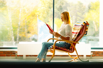 Young attractive woman swinging in a comfortable rocking chair reading her favorite book. Good pastime. Photo with empty side space