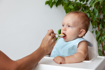 Mother feeds funny happy blond baby in blue bib from a spoon