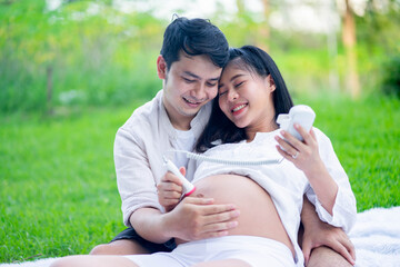 Happy Asian couple pregnant woman and husband using fetal doppler device for listening hear baby...