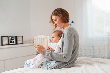 Mother holding a baby and using a smartphone. Side view. White interior. Concept of motherhood and smartphone use