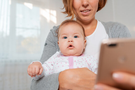 Close-up portrait of a baby sitting in the arms of a mother who uses a smartphone. Bottom view. The smartphone is blurred in the foreground