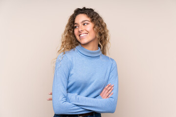 Young blonde woman with curly hair wearing a turtleneck sweater isolated on beige background looking side