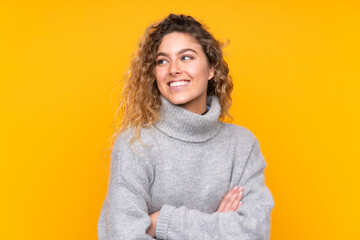 Young blonde woman with curly hair wearing a turtleneck sweater isolated on yellow background...