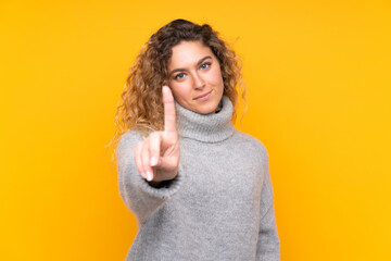 Young blonde woman with curly hair wearing a turtleneck sweater isolated on yellow background showing and lifting a finger