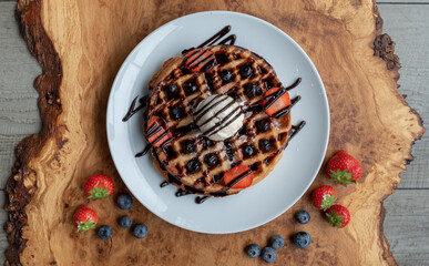 Vegan waffle with vanilla icecream, chocolate sauce, blueberries and strawberries. Photographed from above on a wooden platter.
