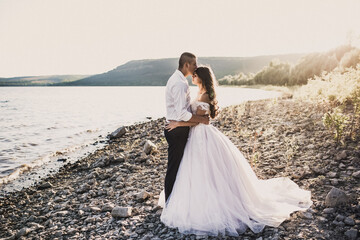 groom and bride in wedding dress stand in embrace on rocky bank near river in summer outdoors In...