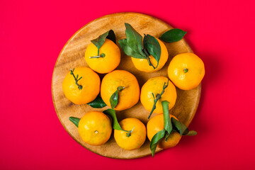 Fresh clementines with leaves on a round board over a red background, top view