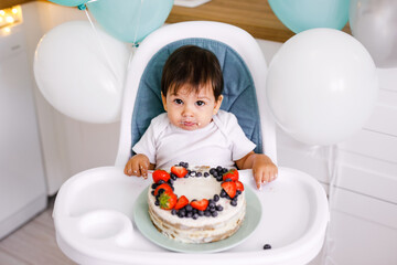 Little baby boy sitting in high chair in white kitchen and tasting first year cake with fruits on background with balloons.