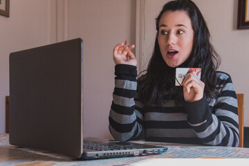 Una mujer joven usa un ordenador portátil para realizar compras por internet mientras sostiene una tarjeta de crédito con una expresión de sorpresa - obrazy, fototapety, plakaty