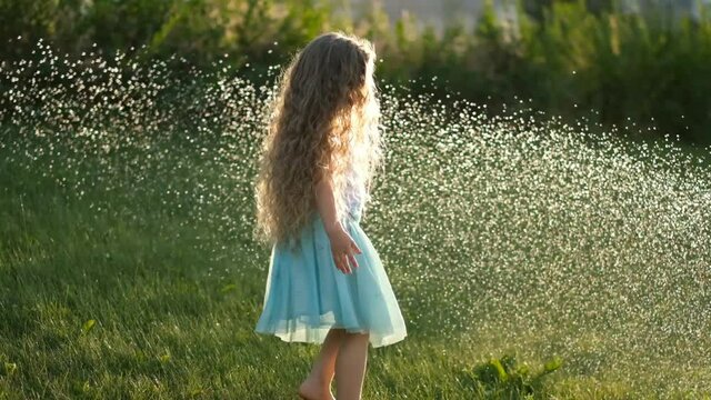 A little girl is having fun on the green lawn, which is poured with a stream of water in summer. Cooling in hot summer under running water on the lawn.