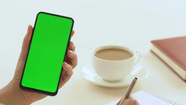Close-up shot of green screen template smartphone in female hands at home offise, girl is watcing content, makes notes in a notebook. Modent technology and information concept.