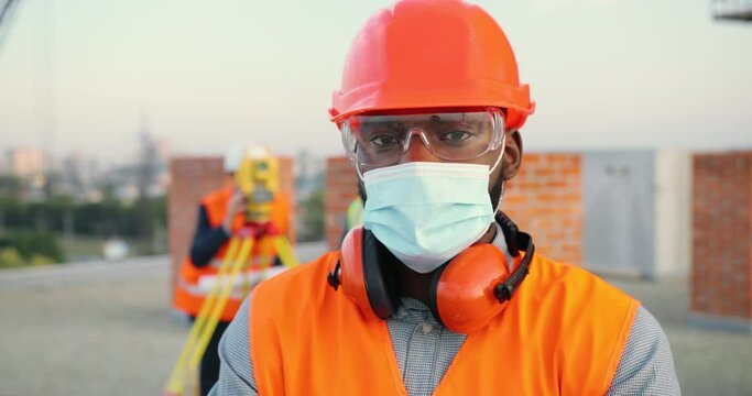 Portrait of African American young man builder in medical mask and casque looking at camera and standing at construction site. Handsome male constructor or architector working outside at building.