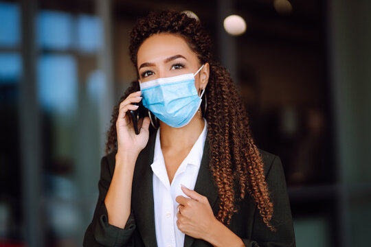 Beautiful serious business woman  in protective medical mask standing near office and talking on phone. Safety during coronavirus pandemic, epidemic covid-19. 