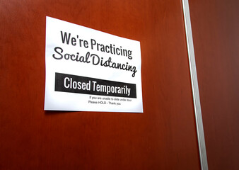Sign on brown office door stating We’re Practicing Social Distancing - Closed Temporarily during the Coronavirus Pandemic