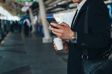Unrecognizable business man holding a disposable coffee cup and using phone at train station