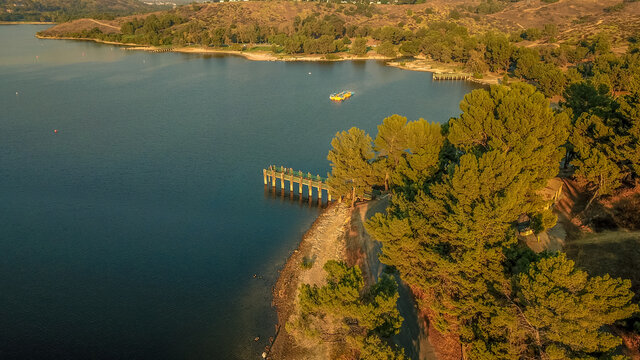a stunning aerial shot of the still blue waters, the pier, the lush green trees and majestic mountain ranges at Puddingstone Lake in San Dimas California