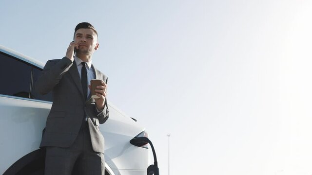 Young successful businessman in suit texts on smartphone while his electric car is charging on background. Side view of white luxury car.