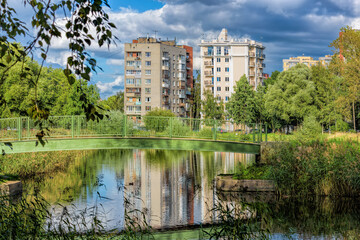 Public city park with pond and reeds and multi-storey dwelling buildings behind under blue sky with amazing clouds at sunny autumn day