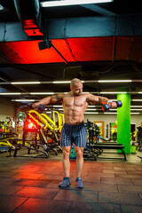 A strong, adult, fit, muscular male trainer poses for a photo shoot in a sports hall under the spotlights in sportswear, demonstrates his muscles and confidently holds dumbbells.