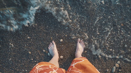 A young man stands on a stone beach and the wave covers his legs. Top view of the legs.