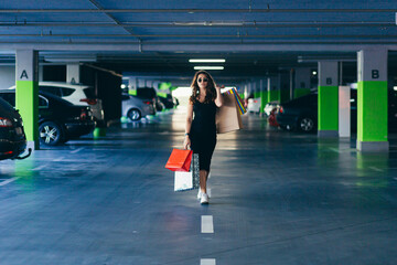 Cheerful lady in stylish black dress carrying on colorful shopping bags in a parking lot near a car. Girl after shopping. Female, caucasian. Attractive brunette looks satisfied after shopping at mall