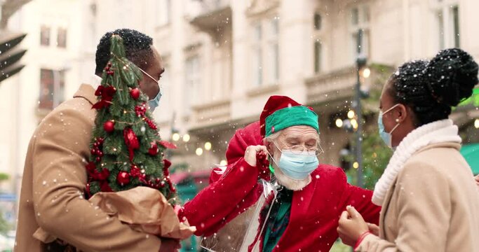Portrait of happy African American man and woman with xmas presents standing outdoors and chatting while jld Caucasian Santa Clause in mask giving wrapped gift to female. Christmas miracle concept