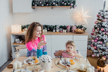 Mom and daughter sitting in the kitchen ha table. Girl playing with Cutlery. the kitchen is decorated for New year and Christmas.