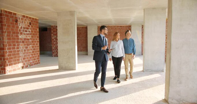 Caucasian male real-estate agent showing apartment at constructing phase to married couple of customers in and talking Wife and husband buying new home flat at building site. Discussion.