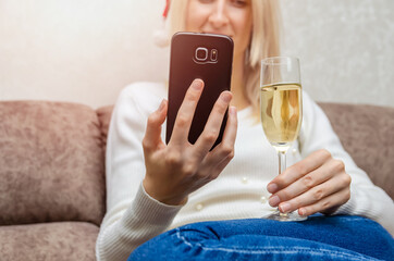 Young beautiful blonde woman in a Santa hat in a white sweater with a glass of champagne smiles and communicates via mobile phone. Concept of Christmas, New year, online communication. Selective focus