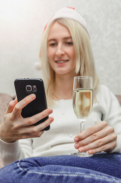 Young beautiful blonde woman in a Santa hat in a white sweater with a glass of champagne smiles and communicates via mobile phone. Concept of Christmas, New year, online communication. Vertical photo