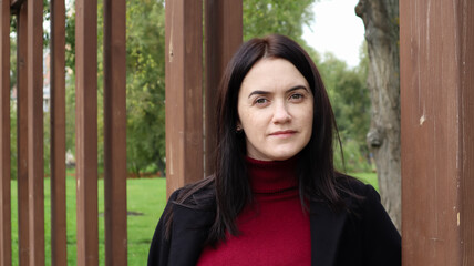 Portrait of a beautiful caucasian woman wearing a black coat and red sweater while walking outdoors in a city park with a blurred background. A woman is looking at the camera on a sunny day.