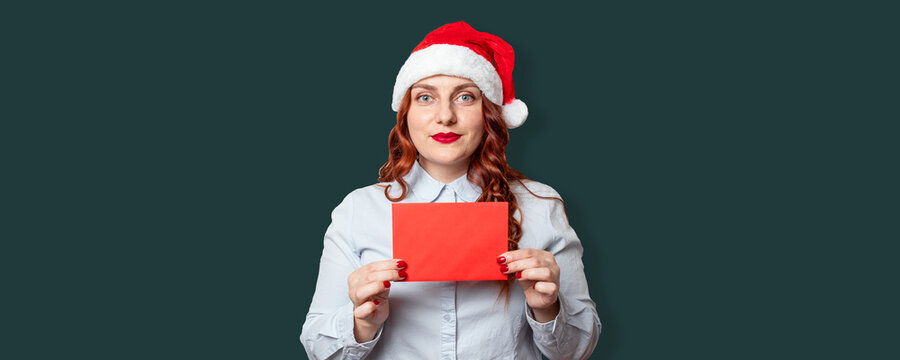 Portrait Of Cheerful Girl In A Santa Hat Holding A Red Wishlist Letter Mockup On Dark Green Wall Background. New Year Celebration Banner, Copy Space.