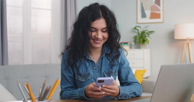 Charming Caucasian woman sitting at table and using smartphone in home office. Happy adult lady texting message and chatting with friends online. Social media, technology concept.