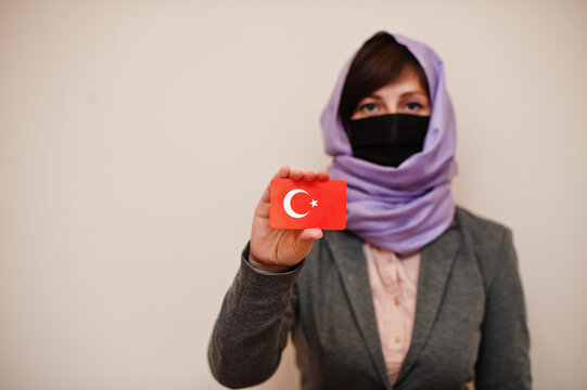 Portrait of young muslim woman wearing formal wear, protect face mask and hijab head scarf, hold Turkey flag card against isolated background. Coronavirus country concept.