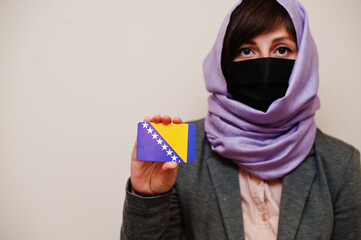 Portrait of young muslim woman wearing formal wear, protect face mask and hijab head scarf, hold Bosnia and Herzegovina flag card against isolated background. Coronavirus country concept.
