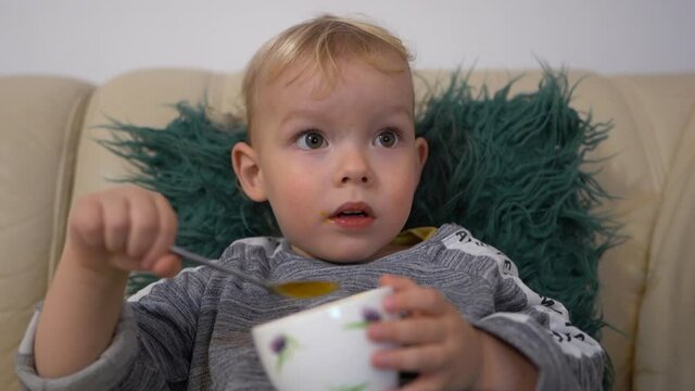 Close indoor portrait of a blond three-year-old. Boy watching cartoons and eating mango ice cream. Happy childhood, leisure child concept