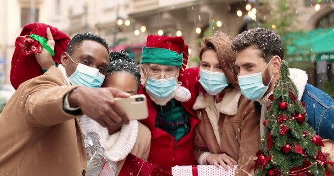 Old Santa Claus hugging African American and Caucasian young people in masks while taking selfie photo on smartphone. Mixed-races friends take pictures with Santa Clause on street. Christmas spirit
