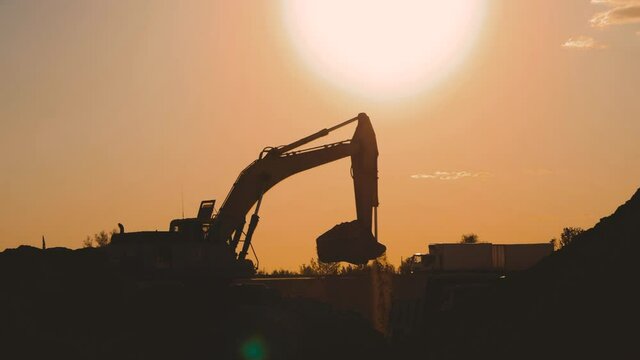 Silhouette of an excavator that loads sand into a truck at sunset. Concept construction and heavy industry, machine will be used in heavy industry business. Slow motion footage.