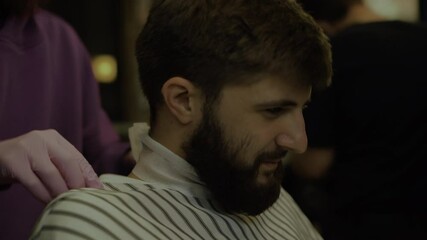 bearded man sitting in a barbershop. Fix the cape for the client