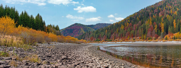 Calm lake with low water - round stones at shore visible, autumn coloured coniferous trees on other side, blue sky above