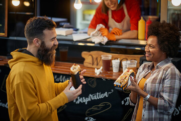 female employee in fast food service smiling, looking at  multiethnic couple in love eating sandwiches