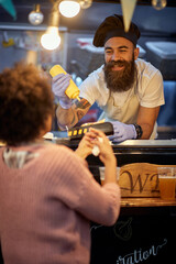 male employee in fast food service smiling and talking to a customer while adding mustard in...