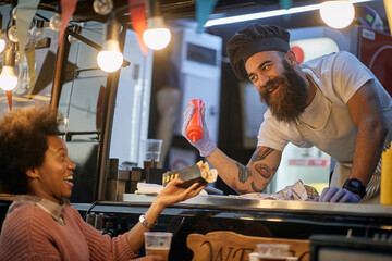 young beardy caucasian employee in fast food adding a ketchup in a sandwich to a female afro-american customer, joking