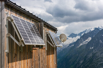 PV solar panels and satellite dish antenna at the wall of a wooden building with snowy mountains in...