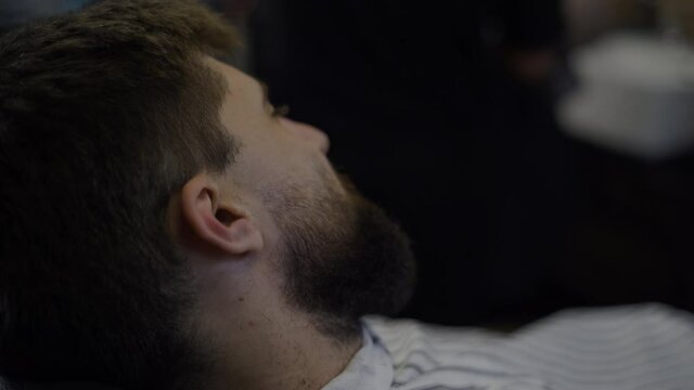 A bearded man trims his beard in a barber shop. Close up of hands of hairdresser working with trimer