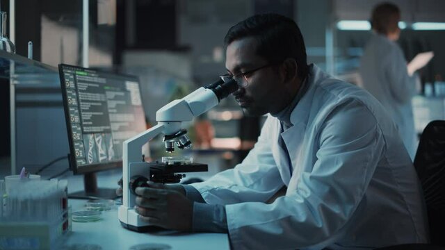 Medical Research Scientist Conducts DNA Experiments Under a Microscope and Writes Results on Desktop Computer in a Biological Science Laboratory. Diverse Multiethnic Lab Engineer in White Coat.