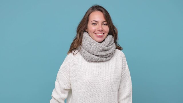 Smiling cheerful young brunette woman in white sweater scarf isolated on pastel blue background in studio. Healthy lifestyle ill sick treatment cold season concept. Looking camera showing thumbs up