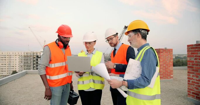 Multi ethnic males and female builders with draft plan of building and laptop computer talking on constructing site. Construction workers and investor, mixed-races men and woman constructors working.