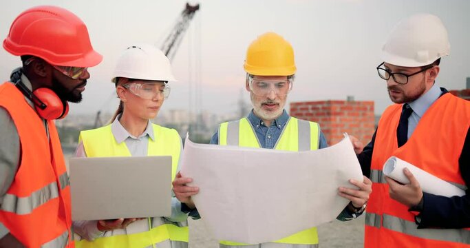 Caucasian and African American males and female builders with draft plan of building and laptop computer talking on constructing site. Architector and investor, men and woman constructors working.