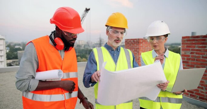 Caucasian and African American males and female builders, architectors and engineers with draft plan of building and tablet talking on constructing site. Multi ethnic men and woman constructors.